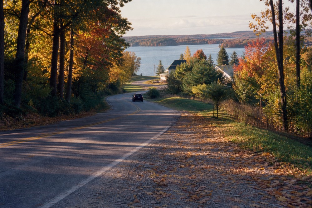 A two-lane road curving through dense autumn hardwoods toward a lake