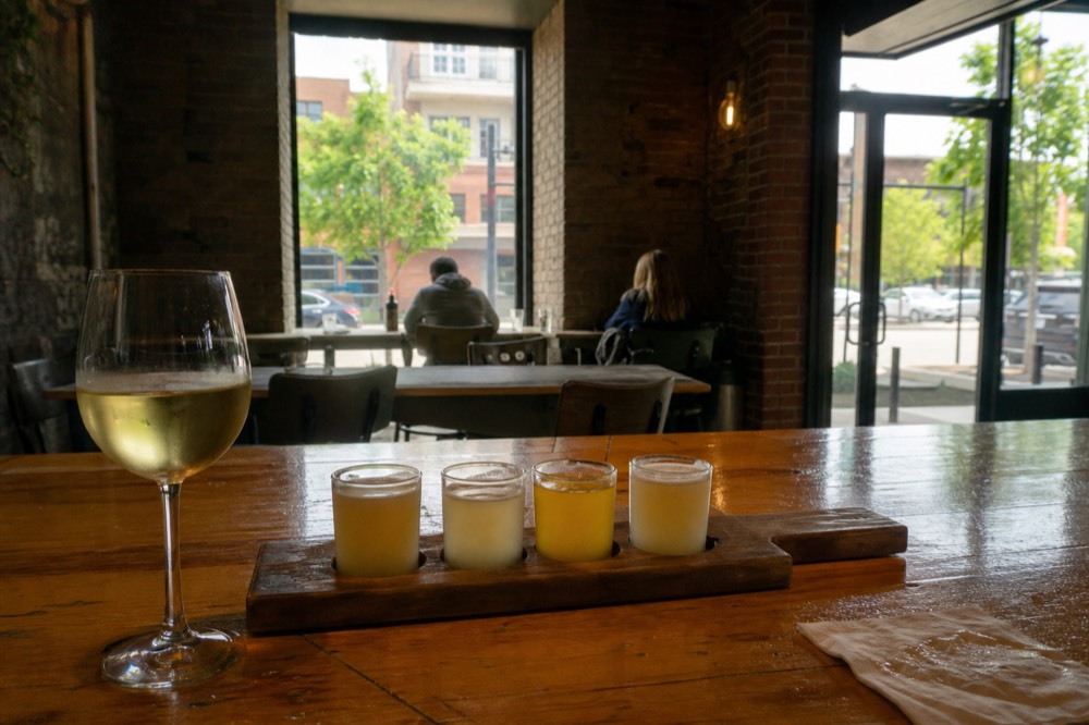 A flight of beers beside a glass of white wine on a wooden bar