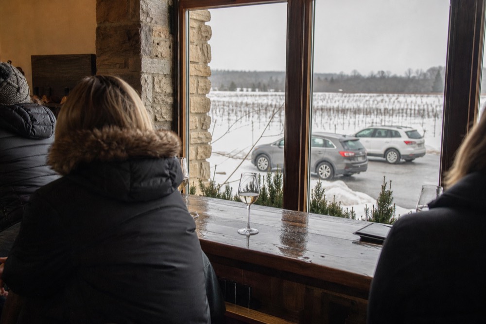 Guests in winter coats tasting wine at an indoor bar with snowy views